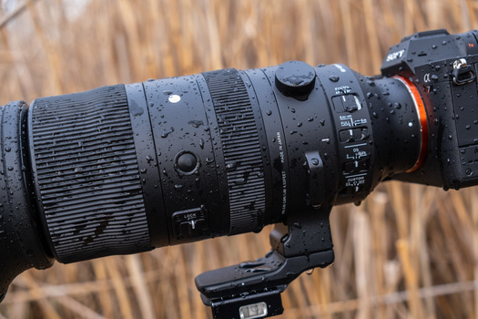 Telephoto lens mounted on camera body, covered in water droplets, with blurred foliage background.