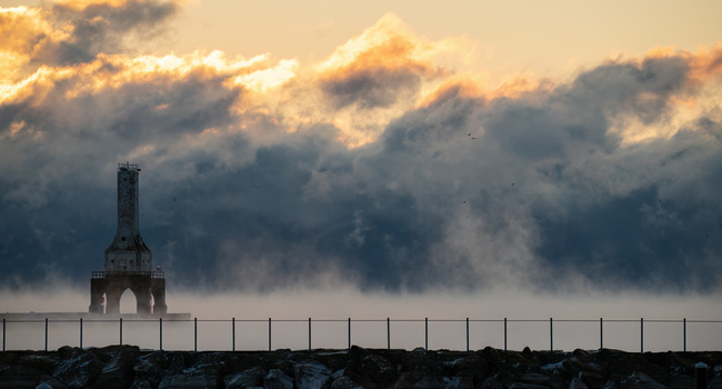 Silhouetted lighthouse structure against dramatic golden and grey storm clouds at dusk.