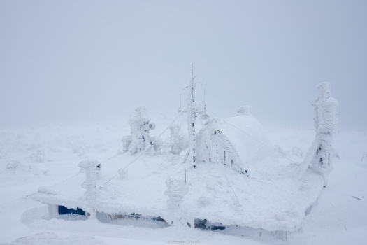 Snow-covered radio transmission towers and equipment on a mountainous landscape during whiteout conditions.