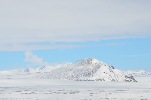 Snow-covered volcanic cone rising from a frozen landscape under clear blue sky.