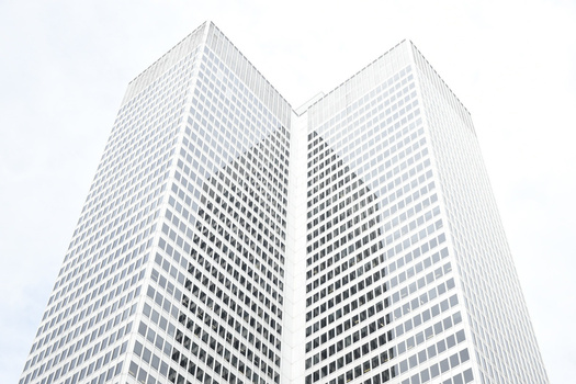 Two modern high-rise buildings with repetitive window patterns converge toward the sky against a light background.