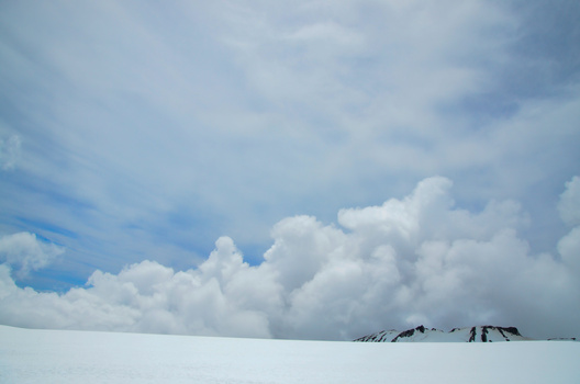 Minimalist snowy landscape with distant structures under dramatic cumulus clouds and blue sky.