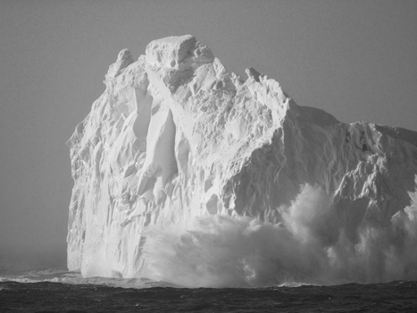Massive iceberg with towering white cliffs and cascading water spray colliding with dark ocean waves.