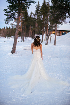 Bride in flowing white gown standing in deep snow with evergreen forest and cabin in background.