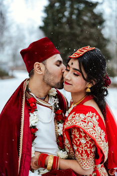 Couple in traditional red wedding attire embrace in falling snow with evergreen trees blurred in background.