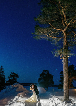 Bride in white wedding dress standing in snowy landscape beneath illuminated tall pine tree at dusk.
