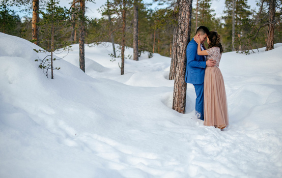 Couple embracing in snow-covered forest with tall pine trees in background.
