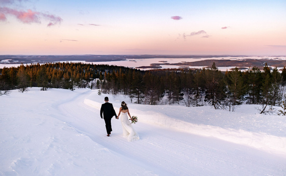 Bride and groom walking across a snowy landscape with forested hills and a frozen lake at dusk.