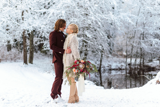Couple embracing in snowy landscape with frost-covered trees and bouquet of flowers.