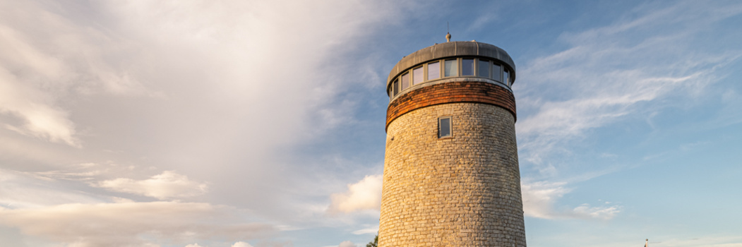 Historic stone lighthouse tower with cylindrical masonry construction and modern glass-topped lantern room against cloudy sky.