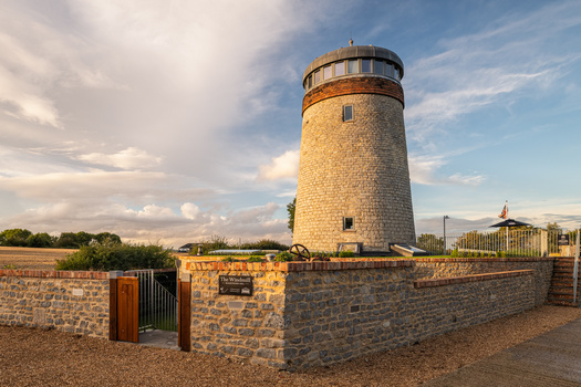 Stone windmill with copper cupola and surrounding brick wall under partly cloudy sky.