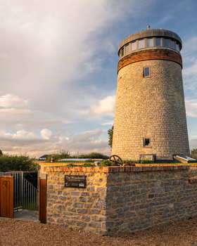 Stone cylindrical tower with rounded top and windows, surrounded by low stone wall and gate under partly cloudy sky.