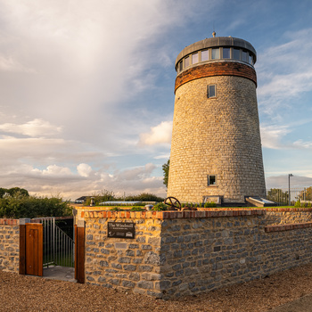 Stone windmill tower with copper-topped lantern room under partly cloudy sky.