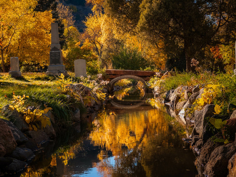 Stone bridge over a reflective stream lined with golden autumn trees in a cemetery.