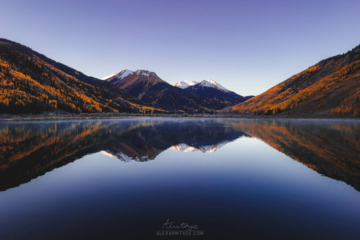 Snow-capped mountain peaks reflected in still lake water, surrounded by golden larch trees in autumn.
