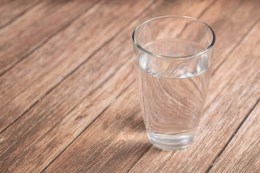 Glass of clear water on wooden surface with soft directional lighting creating subtle shadows.