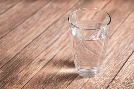Glass of clear water on wooden surface with soft directional sunlight creating shadows.