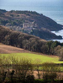 Medieval castle on a forested hillside overlooking a coastal landscape with farmland in the foreground.