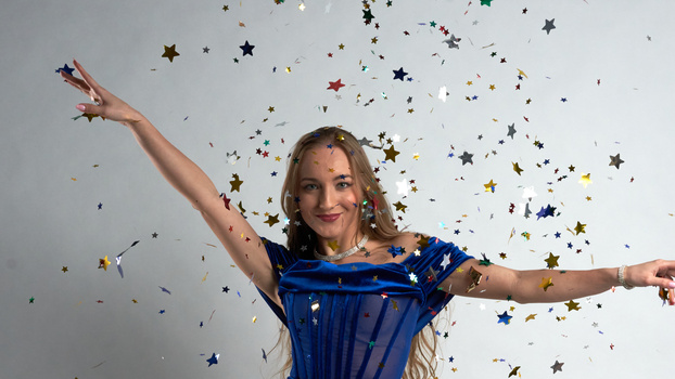 Woman in blue dress with arms raised catching falling gold and multicolored stars against white background.