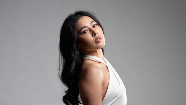 Woman with long dark hair in white halter top against neutral gray background, looking over shoulder.