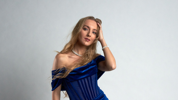 Woman in blue off-shoulder corset dress against neutral background, hand raised to head.