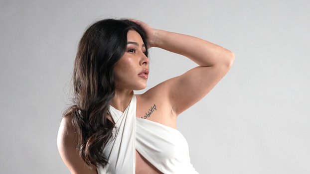 Woman in white draped top with hand raised to head, looking upward against neutral gray background.
