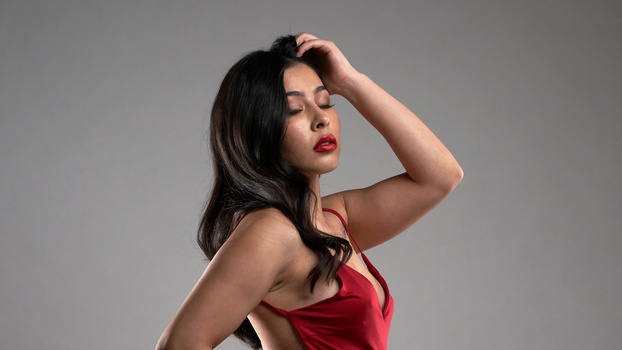 Woman in red tank top with hand to hair, looking down in profile against gray background.