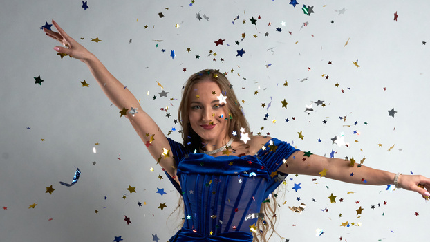Woman in blue dress with arms outstretched amid falling gold and silver star confetti against white background.
