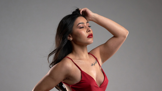 Woman in red dress with hand to head, shot against neutral gray background in profile.