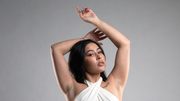 Woman with arms raised above head, looking upward against neutral gray background.