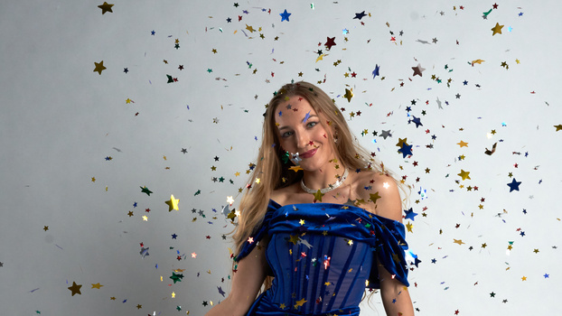 Woman in blue off-shoulder dress surrounded by falling confetti and star-shaped decorations.