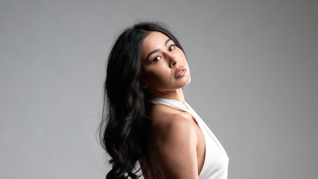 Woman with long dark hair looking over her shoulder against a neutral gray background.