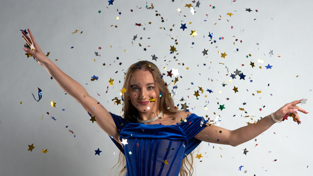 Woman in blue dress with arms raised, surrounded by falling star-shaped confetti.