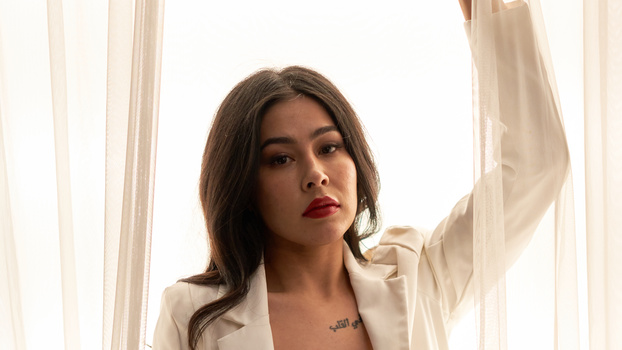 Woman with dark hair and red lipstick gazing upward beside sheer white curtains.
