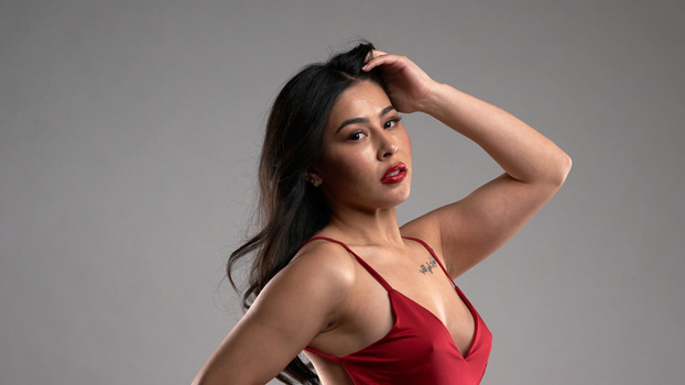 Woman in red camisole posing against neutral gray background with hand in hair.