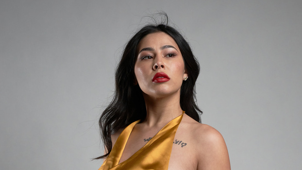 Woman with long black hair wearing a gold sleeveless top against a neutral gray background.