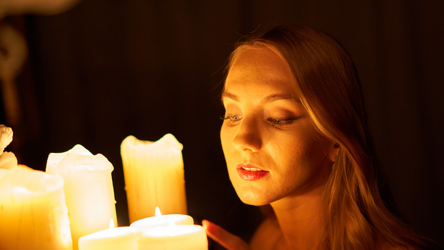 Woman with red lips gazing downward, illuminated by warm candlelight against a dark background.