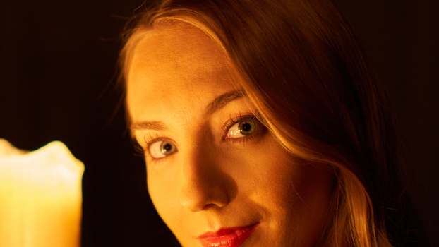 Woman's face illuminated by warm, golden candlelight against a dark background.