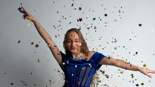Woman in blue dress throwing gold and silver star confetti with both arms raised against neutral background.