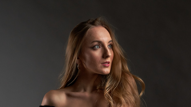 Woman with long brown hair against dark gray background, looking over shoulder.