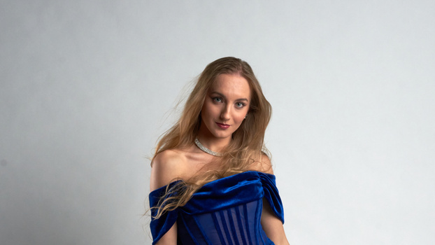 Woman with long brown hair wearing an off-shoulder blue velvet top against a neutral gray background.