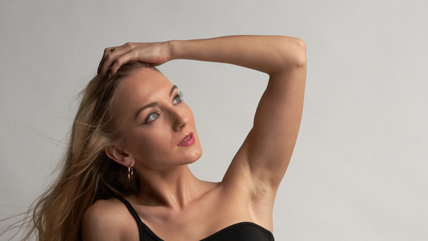 Woman with hand raised to head, looking upward against neutral background.
