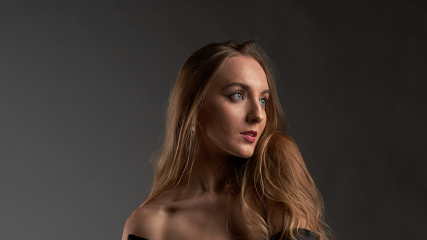 Woman with long brown hair against neutral gray background, looking over shoulder.
