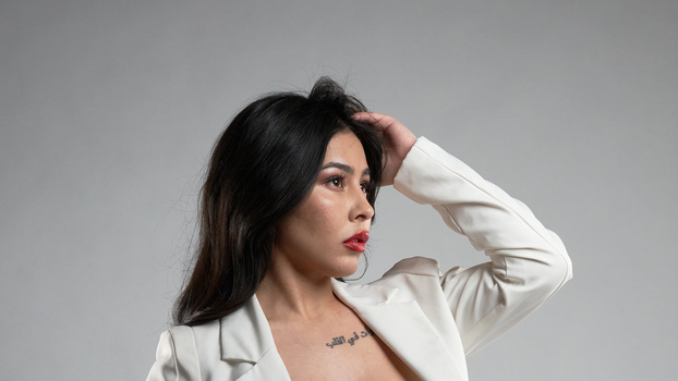 Woman in cream blazer gazing to the side with hand raised to head against neutral gray background.