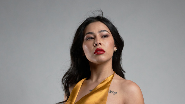 Woman with long dark hair wearing a yellow sleeveless top, photographed against a neutral gray background.