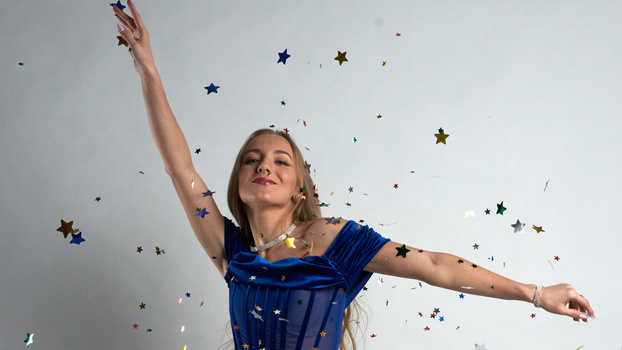 Woman in blue dress with arms raised, surrounded by falling confetti and star decorations against a white background.