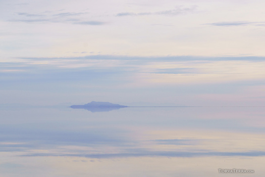 A small island silhouette centered in calm water under a soft, hazy sky at dawn or dusk.