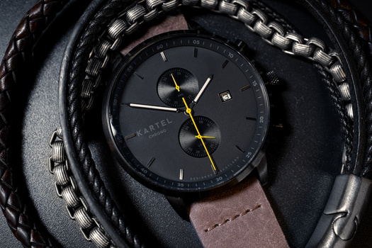 Chronograph watch with dark dial and brown leather strap on textured fabric background.