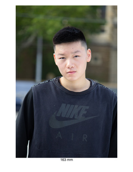 Young man in dark Nike sweatshirt looking directly at camera against blurred outdoor background.