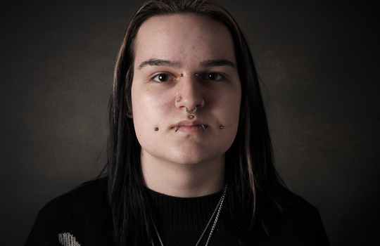 Portrait of a person with long dark hair and facial piercings against a dark background.